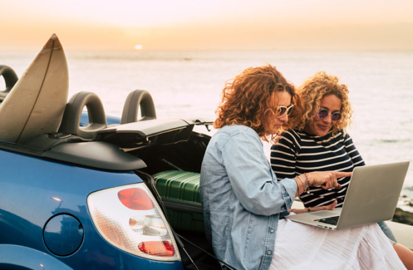 Two women consult a laptop while sitting on the back of their convertible near the ocean